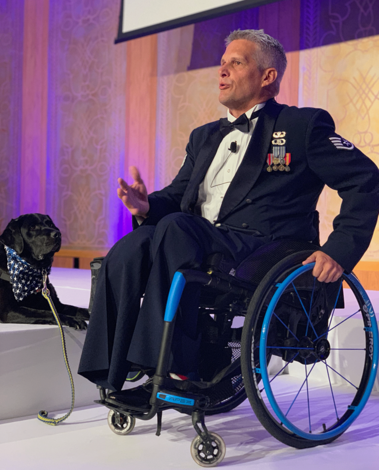 Jason Morgan speaking on stage in Air Force dress uniform with his service dog Yago